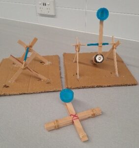 Photo showing a completed trebuchet (right) and catapult (front) built by Levi and Ali. They worked consistently and were the first group to complete their STEM challenge. Also displayed: an incomplete trebuchet by another group.